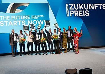 Gewinnerinnen und Gewinner des INTERNORGA Zukunftspreises 2024 jubeln auf der Bühne mit Trophäen, Urkunden und Blumen. Im Hintergrund die Leinwand mit „The Future Starts Now!“