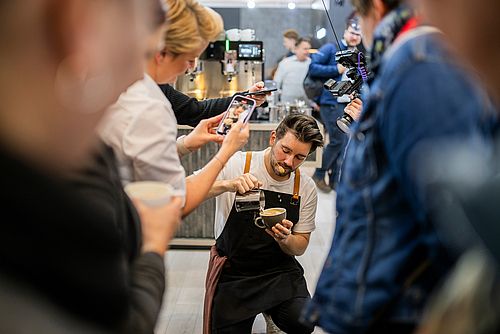 Ein Barista wird beim Latte Art machen von einigen Personen gefilmt.