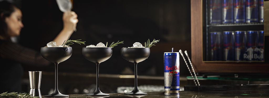 Bartresen mit drei Cocktailgläsern und einer Red Bull Dose, im Hintergrund weitere Red Bull-Dosen und eine Barkeeperin