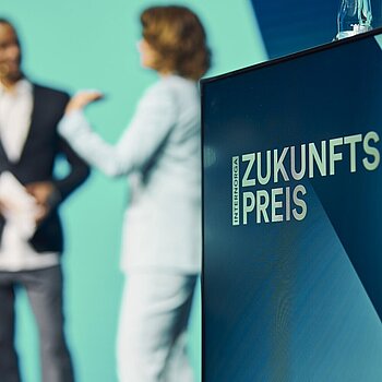 Im Vordergrund ist ein Rednerpult mit der Aufschrift "Zukunftspreis" zu sehen, während im Hintergrund unscharf eine Frau in einem hellen Anzug und ein Mann in dunkler Kleidung auf der Bühne miteinander sprechen.