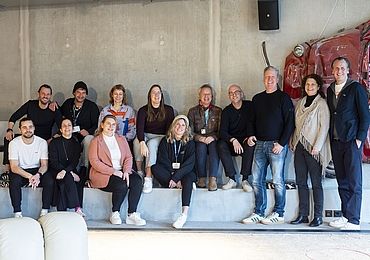 Eine Gruppe von 13 Personen sitzt entspannt auf Stufen in einem modern gestalteten Raum. Sie lächeln in die Kamera, im Hintergrund ist ein in die Wand integriertes, rotes Oldtimer-Frontteil zu sehen.