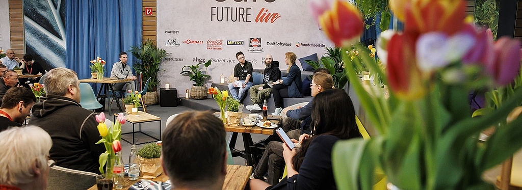 Eine inspirierende Gesprächsrunde im „Café Future live“ auf der INTERNORGA. Das Publikum sitzt an Tischen mit Blumen und Getränken, während auf der Bühne mehrere Personen über Branchenthemen diskutieren.