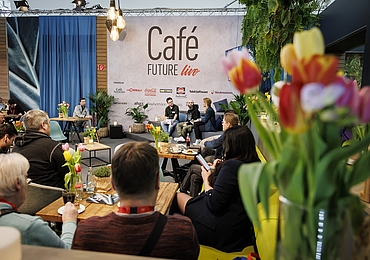 Eine inspirierende Gesprächsrunde im „Café Future live“ auf der INTERNORGA. Das Publikum sitzt an Tischen mit Blumen und Getränken, während auf der Bühne mehrere Personen über Branchenthemen diskutieren.