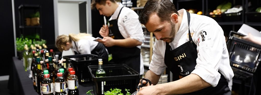 Drei Köche arbeiten während des Next Chef Awards an einer Küchenstation mit frischen Kräutern und Zutaten. Ein Koch schneidet Kräuter im Vordergrund.