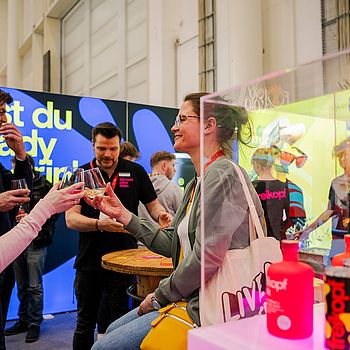 Eine fröhliche Szene auf einer Messe, bei der zwei Frauen mit Gläsern anstoßen. Im Hintergrund sind weitere Personen an einem Stand mit einer farbenfrohen Kulisse und der Aufschrift „Bist du ready?“ zu sehen.