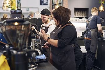 zenenbild aus einem Café mit mehreren Baristas, die an einer Kaffeestation arbeiten. Sie tragen Schürzen und wirken konzentriert und gut gelaunt, während sie Kaffee zubereiten.