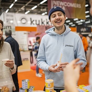 Zwei Messebesucher probieren eine Kostprobe an einem Stand in der „Newcomers Area“. Ein junger Mann mit hellblauem Hoodie und Mütze lacht und hält ein Probierbecherchen, während eine Frau in einem hellen Pullover ebenfalls eine Probe kostet.