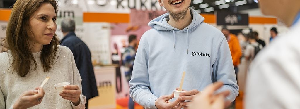 Zwei Messebesucher probieren eine Kostprobe an einem Stand in der „Newcomers Area“. Ein junger Mann mit hellblauem Hoodie und Mütze lacht und hält ein Probierbecherchen, während eine Frau in einem hellen Pullover ebenfalls eine Probe kostet.