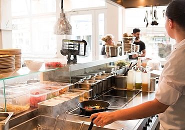 Blick in die Küche eines Restaurants, ein Koch steht mit einer Pfanne am Herd