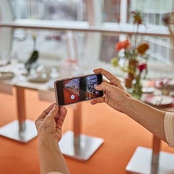 Eine Person fotografiert mit einem Smartphone den „Skywalk“, eine modern gestaltete Fläche mit elegant gedeckten Tischen, floralen Dekorationen und großen Fenstern, die für eine lichtdurchflutete Atmosphäre sorgen.